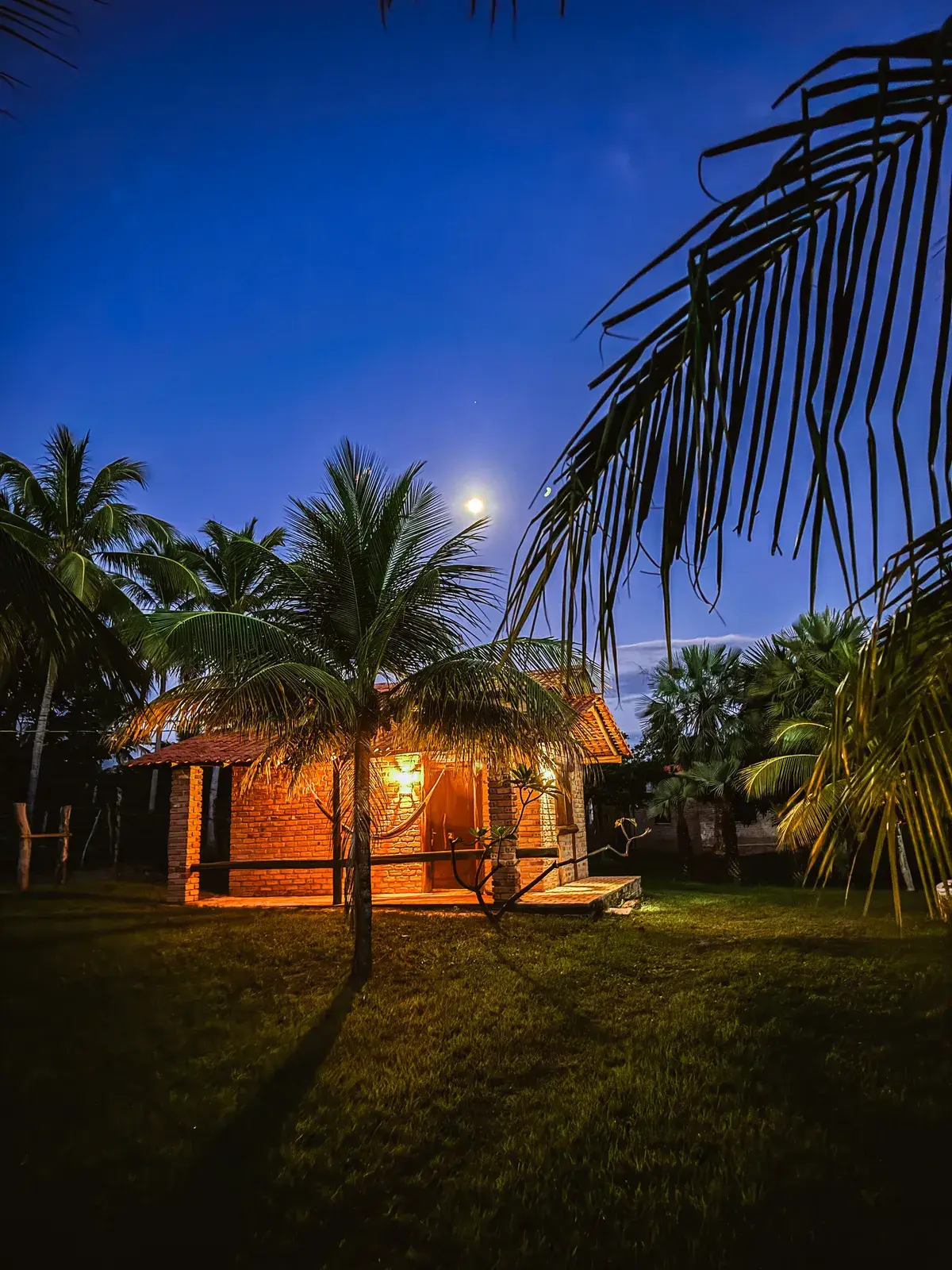 O Rancho ao anoitecer: uma casa de tijolo com a varanda iluminada e uma rede, entre coqueiros, sob a lua