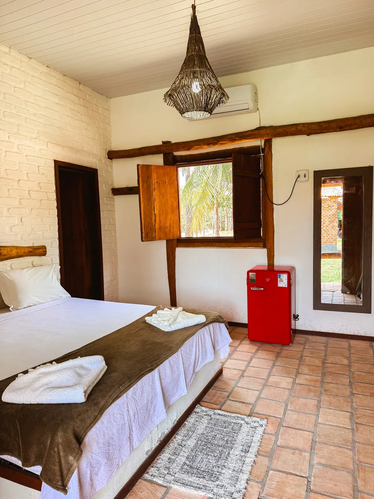 Cama de casal no Bungalow Triplo, com janela de madeira aberta, frigobar vermelho e espelho com moldura de madeira