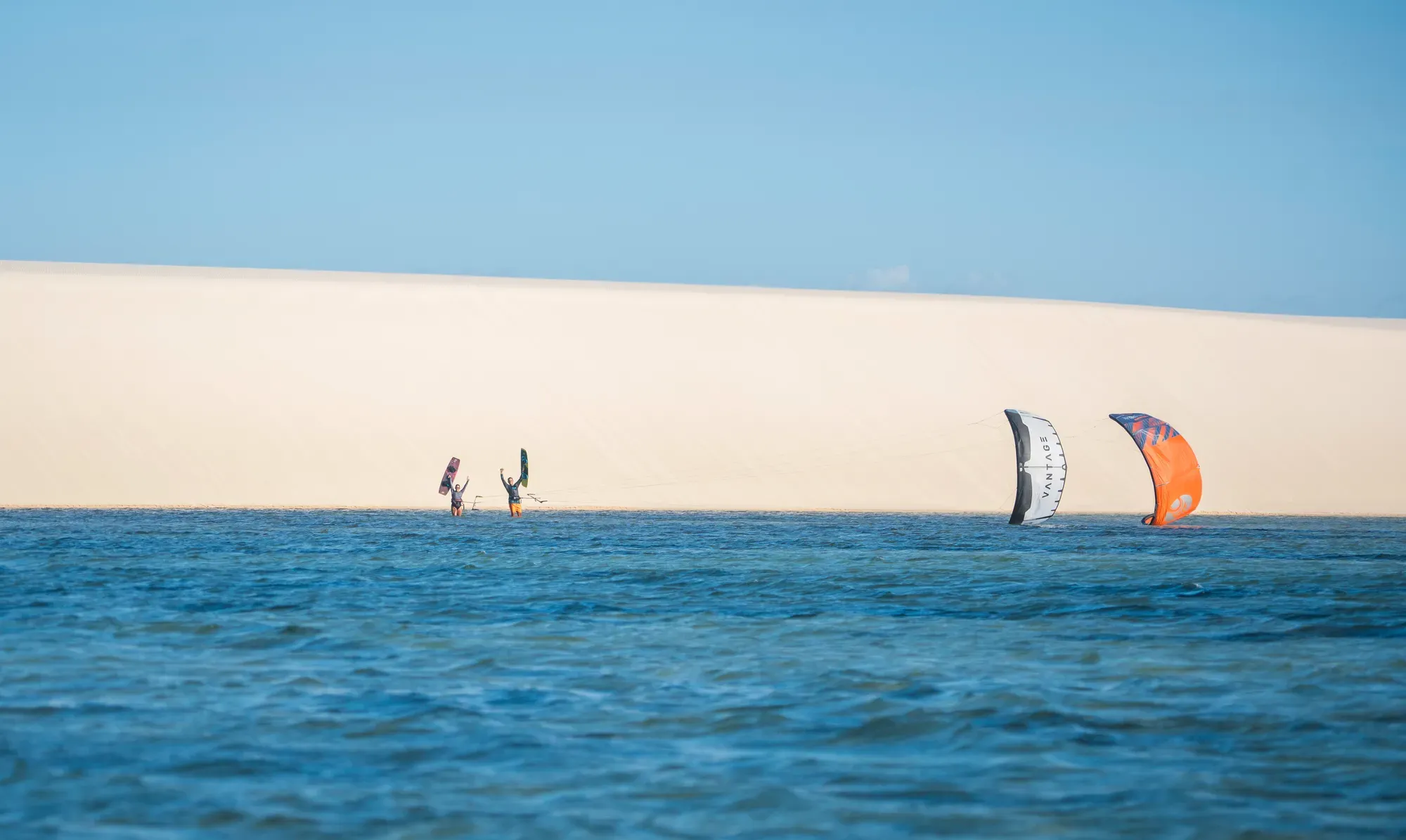 Duna branca e lagoa azul nos Lençóis Maranhenses, com dois kitesurfistas e seus kites na beira da água