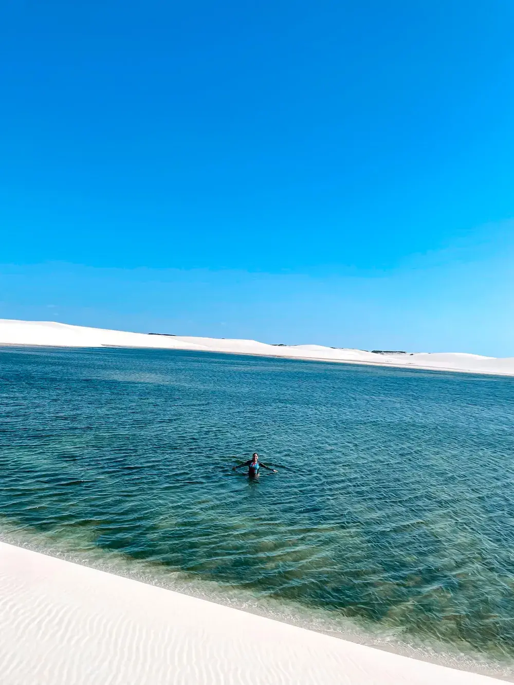 Uma pessoa de braços abertos numa lagoa azul-turquesa, ao lado de uma duna branca
